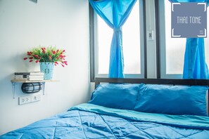 A bedroom with a bed, a window with blue curtains, a wall-mounted shelf with books and a plant, and a wall socket.