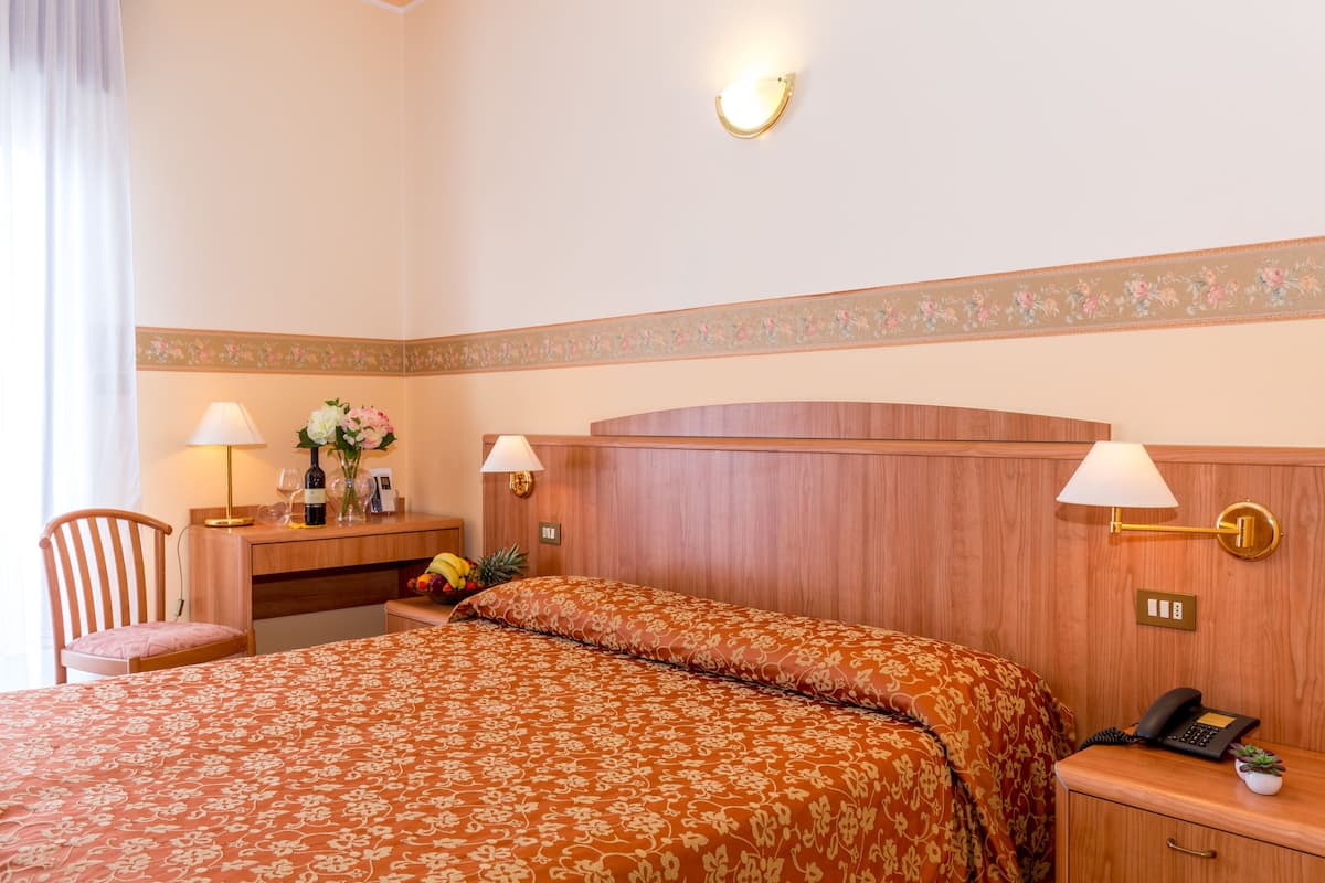 Habitación de hotel con cabecera de madera, cama con colcha estampada, escritorio con lámpara, una silla y una mesita con una planta.