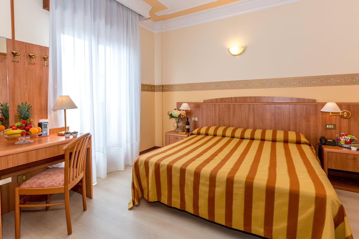 Una habitación de hotel con un escritorio de madera, una silla, una cama con ropa de cama a rayas, una mesita de noche con lámpara y un espejo instalado en la pared.