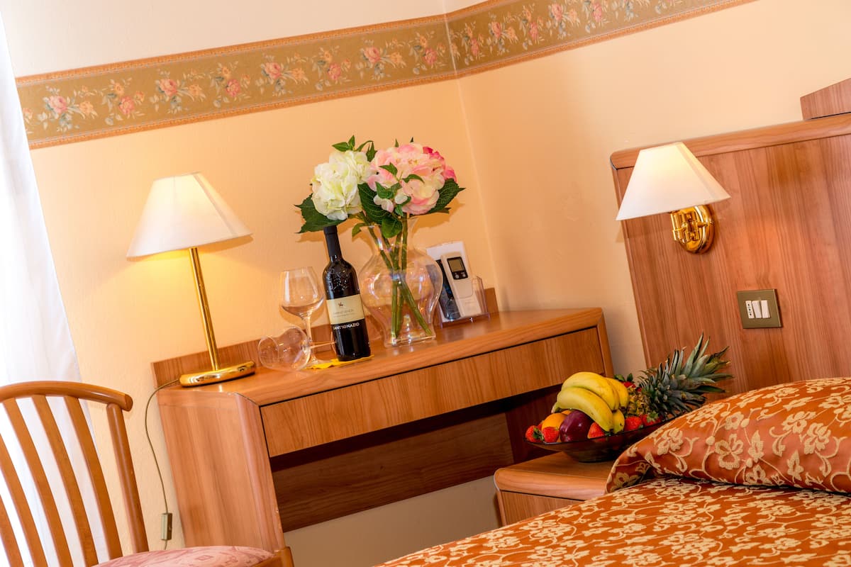 Una habitación de hotel con un escritorio de madera, dos lámparas, un florero con flores, una botella de vino y un frutero en la mesita de noche.
