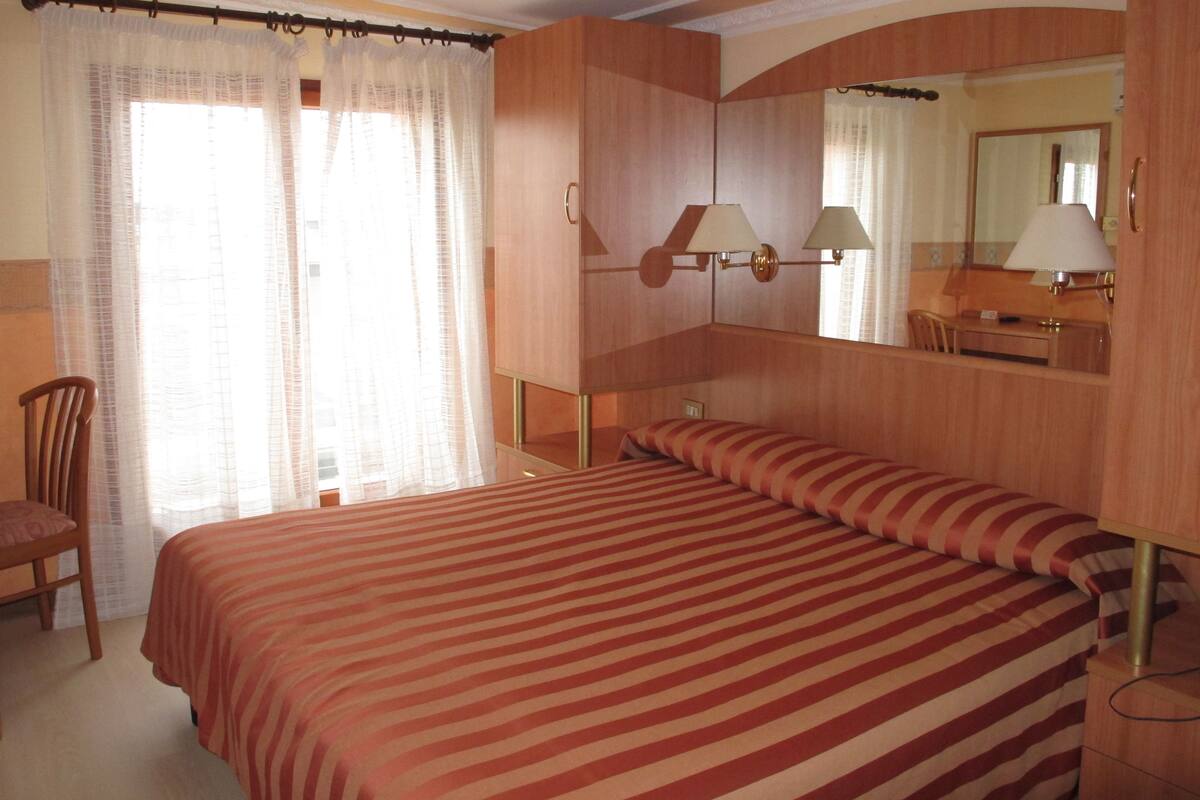 Una habitación de hotel con una cama grande, una silla, un escritorio, un espejo y una ventana con cortinas.