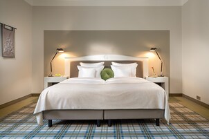 Una cama bien hecha con una colcha blanca, dos lámparas de noche y un almohadón verde. La habitación tiene un suelo a cuadros y una pared lisa con un cuadro enmarcado.
