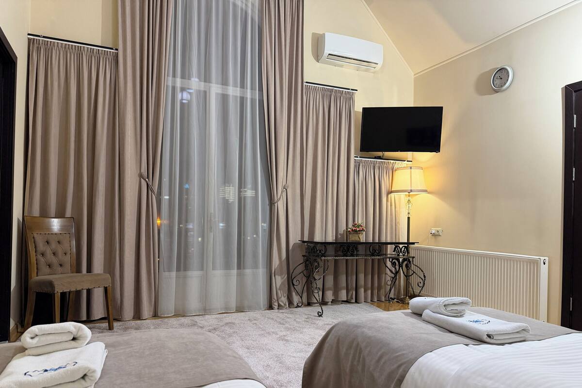 A hotel room with two beds, a chair, a table with a lamp, an air conditioner, a clock, and a view of the city at night.
