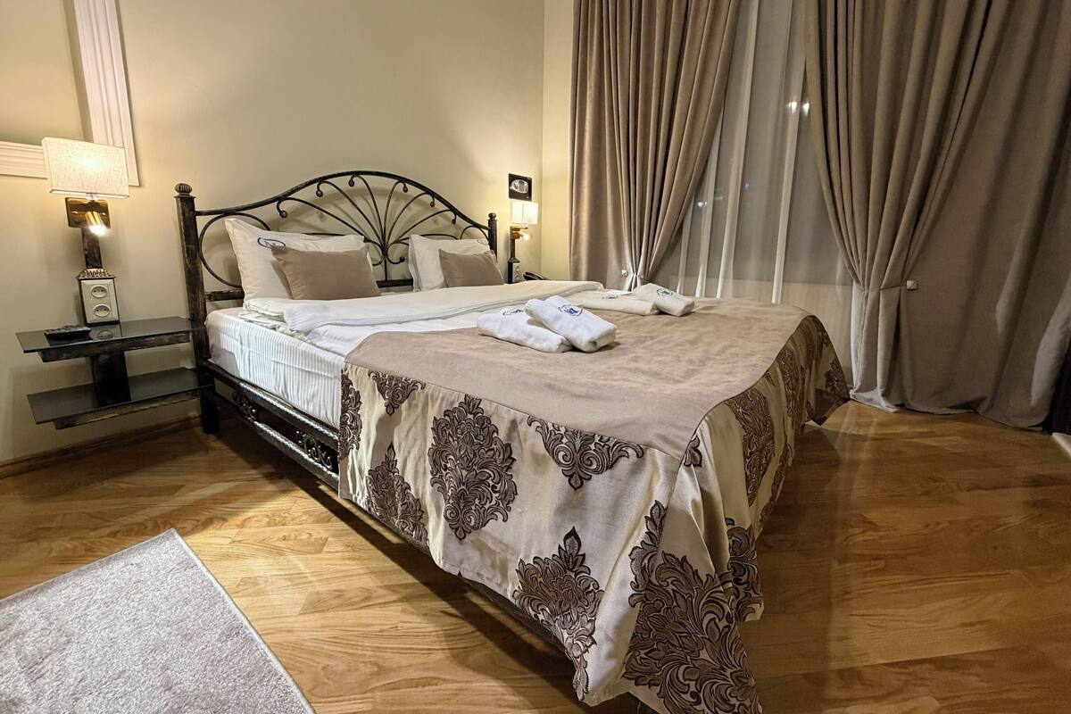 A neatly made bed with a patterned bedspread, a bedside table with a lamp, and a window with curtains.