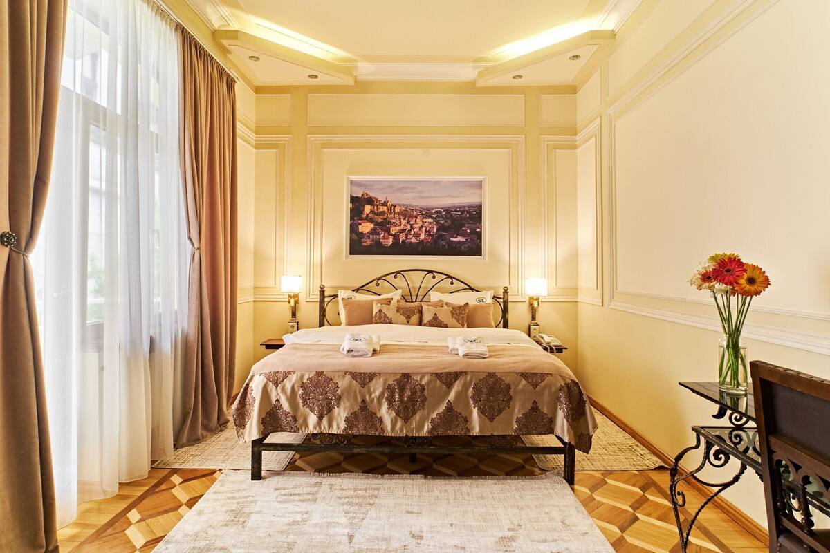 A hotel room with a bed, a painting on the wall, a vase with flowers, and a wooden bench.