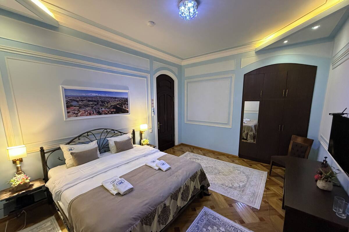 A well-lit bedroom with a large bed, a view of a cityscape, a dark wardrobe, and a desk with a plant.