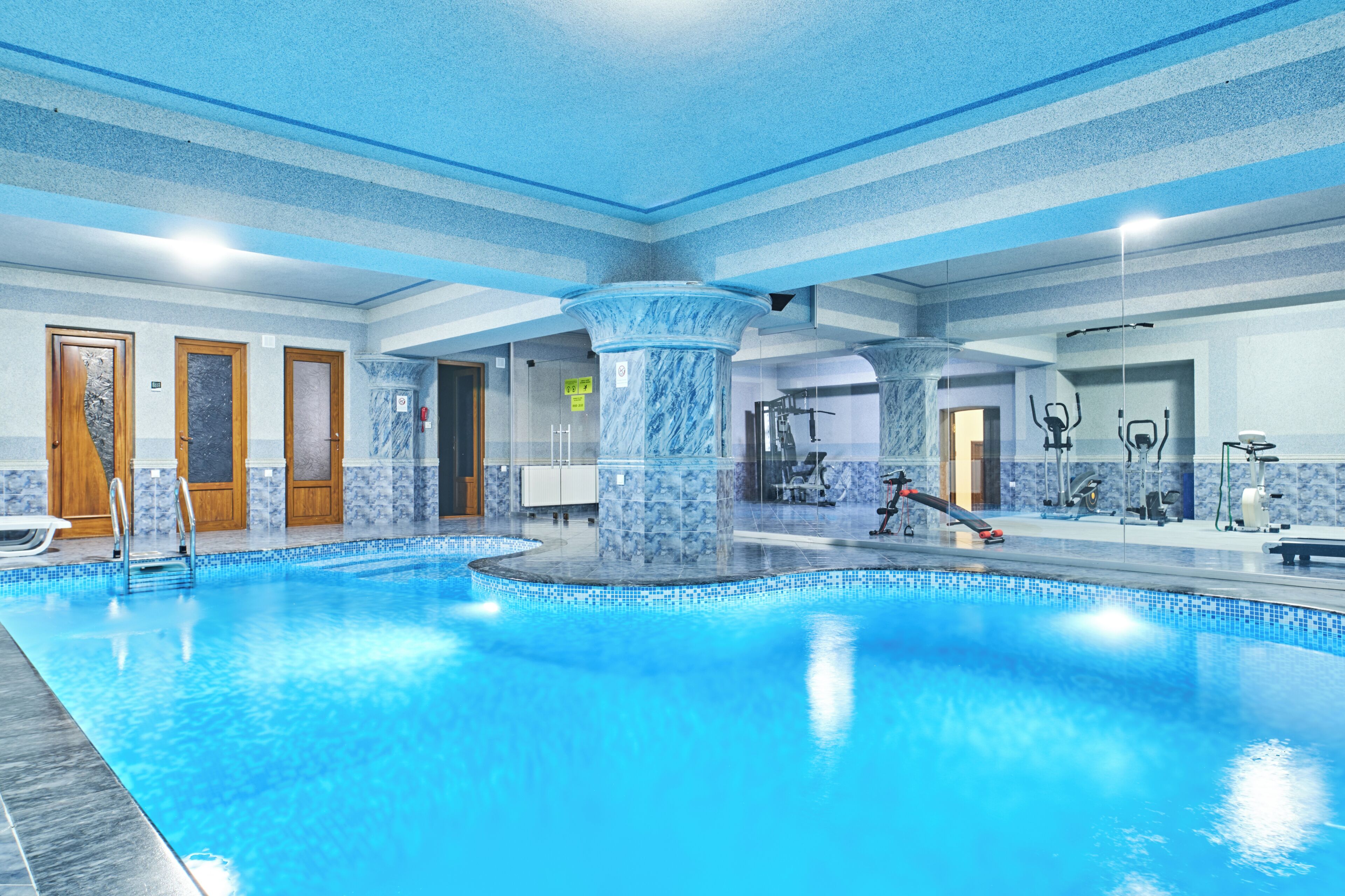 An indoor swimming pool with a blue and white color scheme, surrounded by exercise equipment and a wooden door.