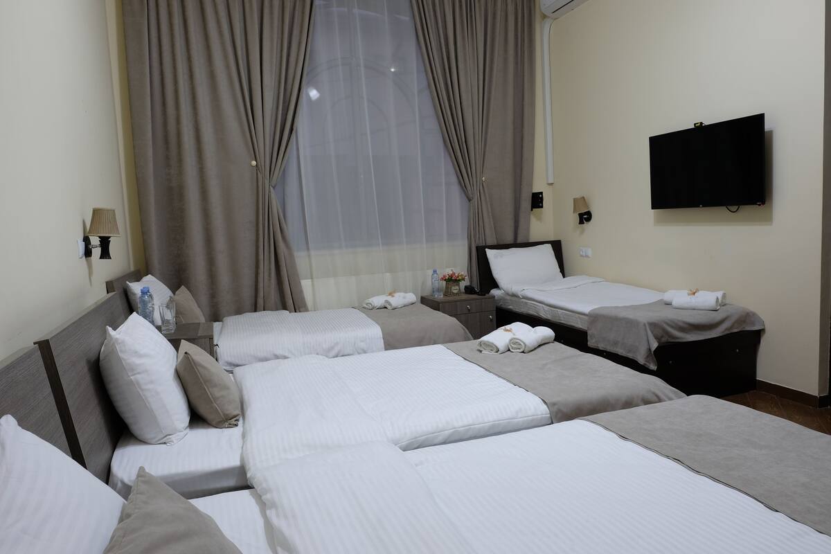A hotel room with three beds, a television, a window with curtains, and a bedside table with a lamp and a bottle.