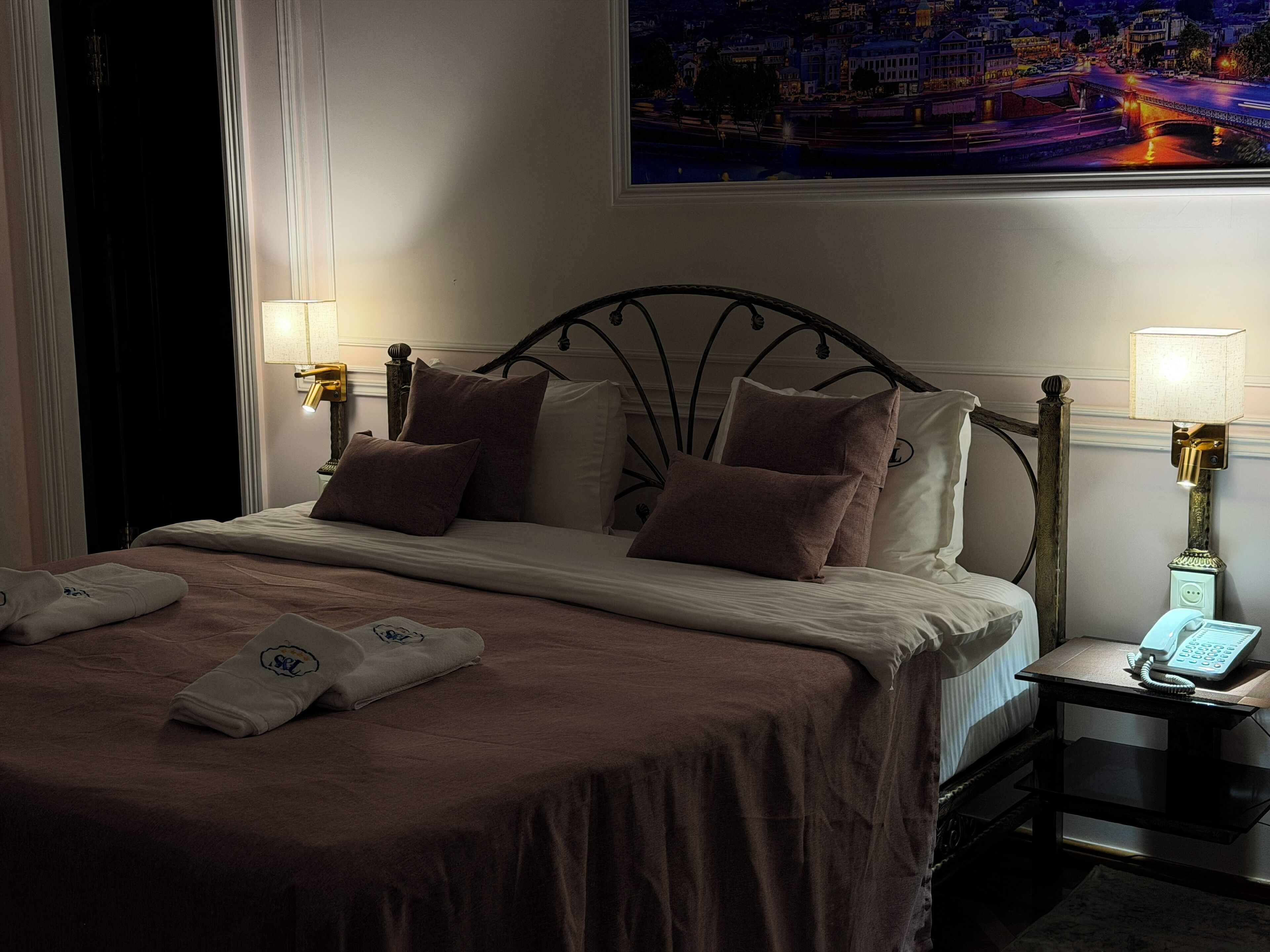 Habitación de hotel con una cama, dos mesitas de noche, una lámpara y vistas de la ciudad en la pared.