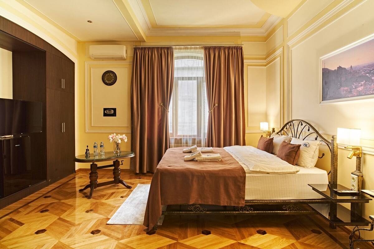 A hotel room with a bed, a desk, a chair, a TV, a window with curtains, a painting on the wall, and a clock.
