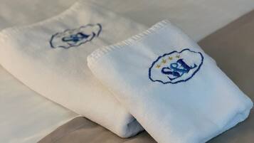 A neatly made bed with white linens and two towels with embroidered logos.