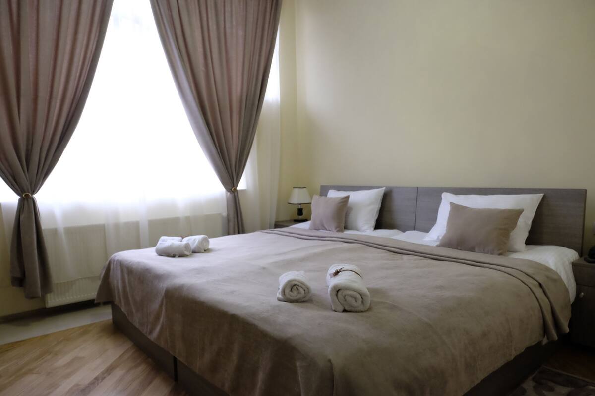 A neatly made bed with a beige bedspread, two rolled towels, and a nightstand with a lamp.
