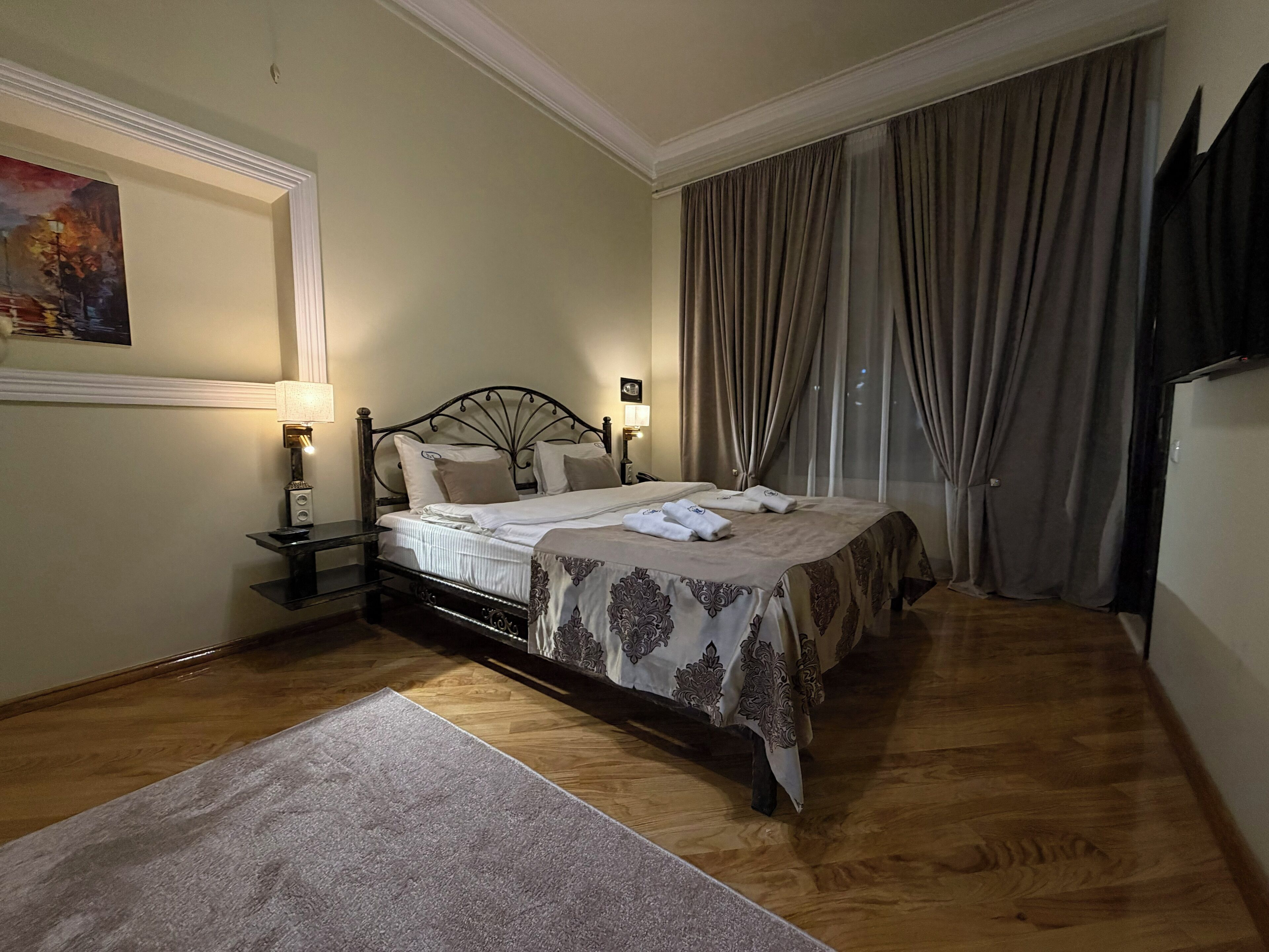Un dormitorio ordenado con cama, mesitas de noche, un ventanal con cortinas y un cuadro en la pared.