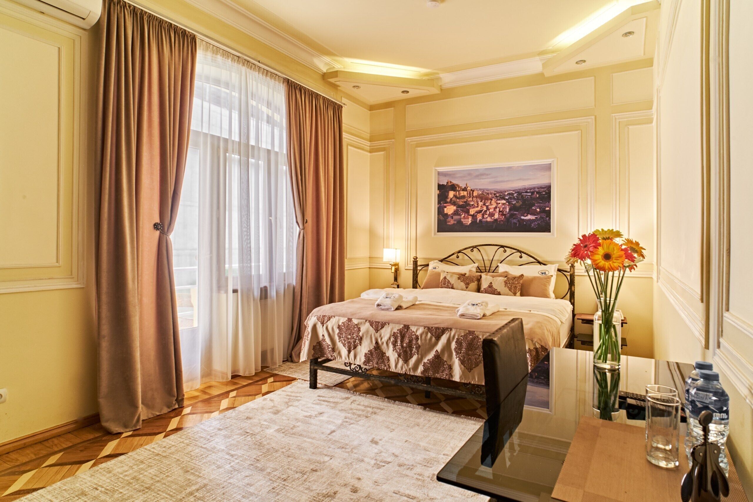 A hotel room with a bed, a desk with a chair, a vase with flowers, a painting on the wall, and a window with curtains.