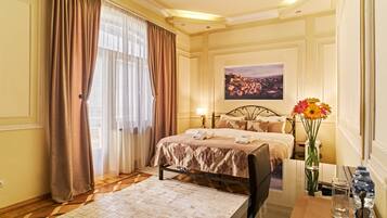 A hotel room with a bed, a desk with a chair, a vase with flowers, a painting on the wall, and a window with curtains.
