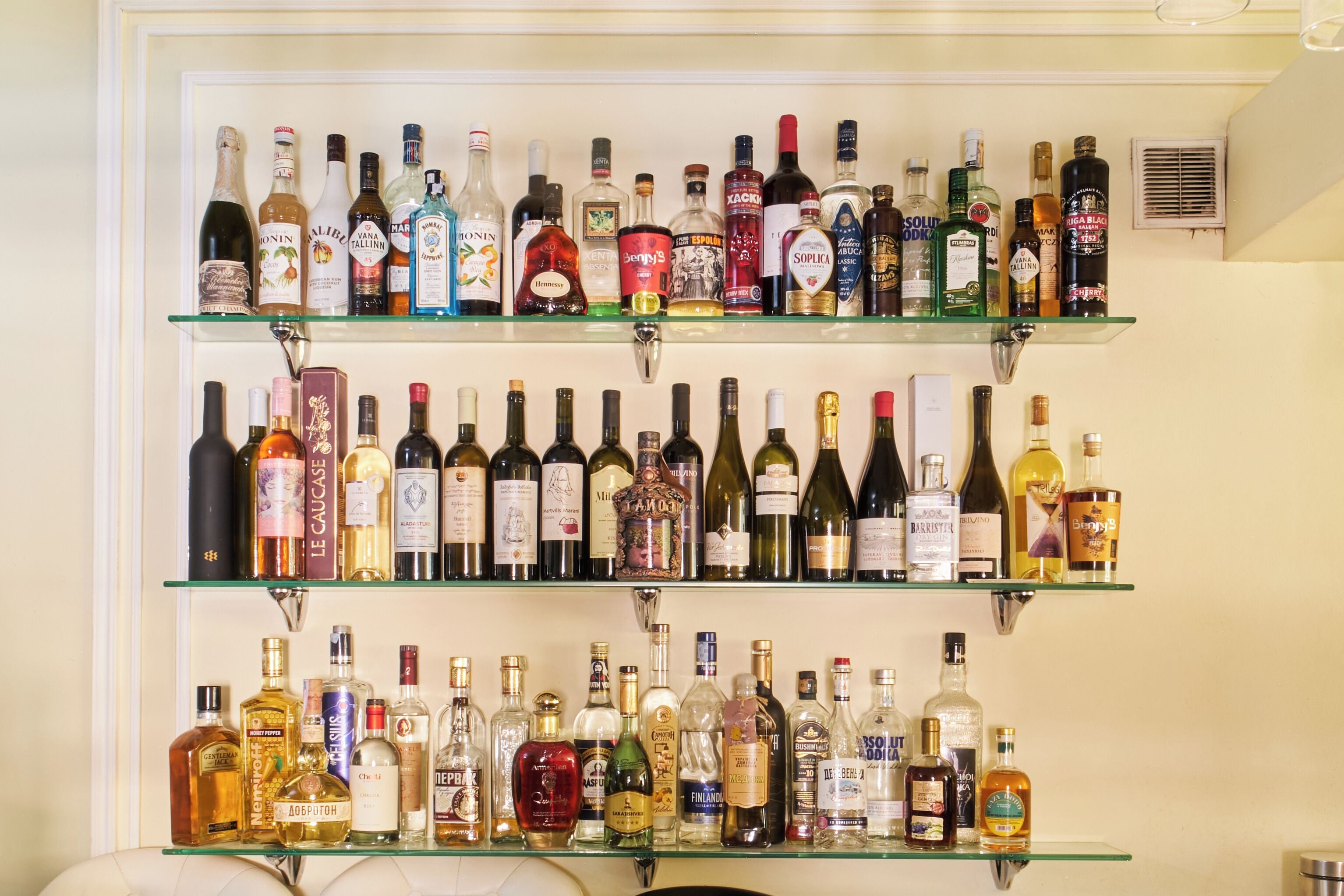 A wall with three shelves of various liquor bottles.