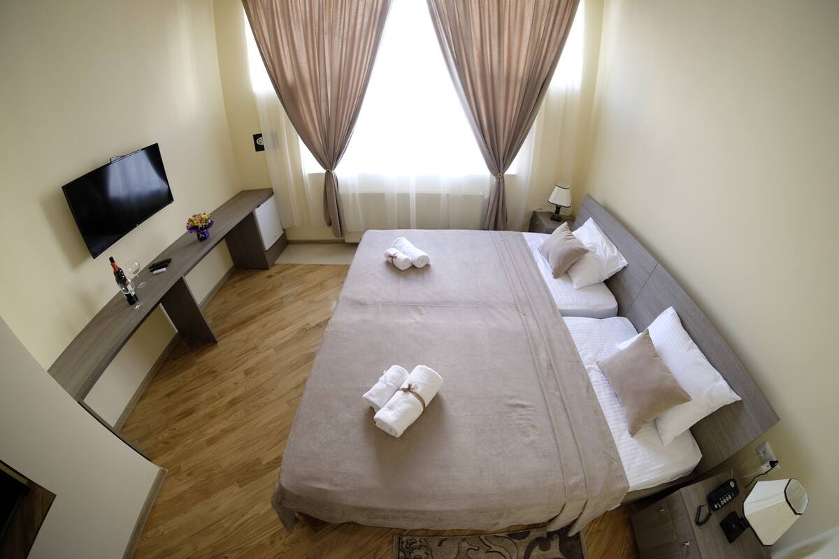 A hotel room with a large bed, a TV, a bedside table with a lamp, and a window with curtains.