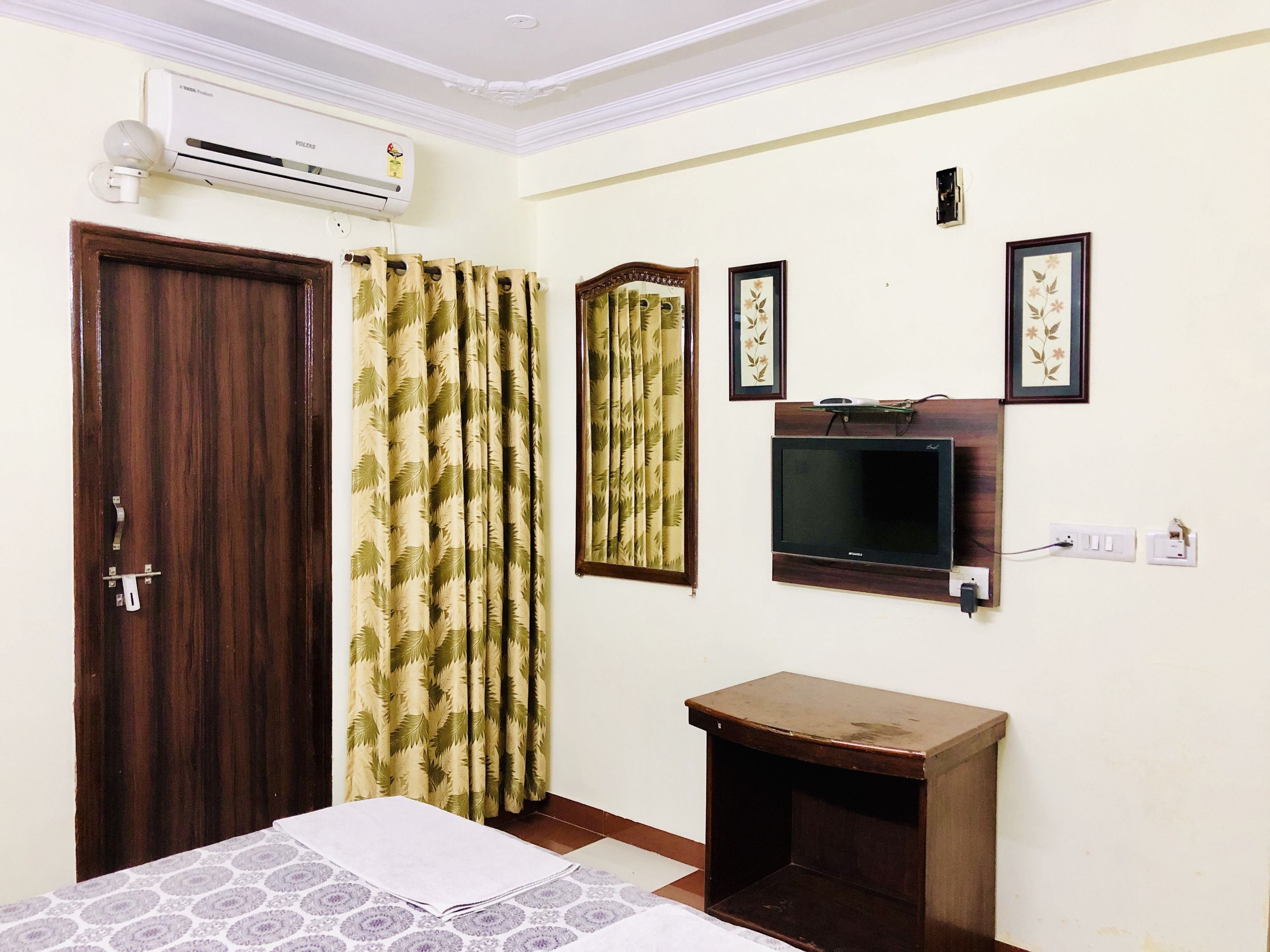 Una habitación con una cama, una mesita de noche de madera, un televisor montado en la pared y una puerta con cortina.