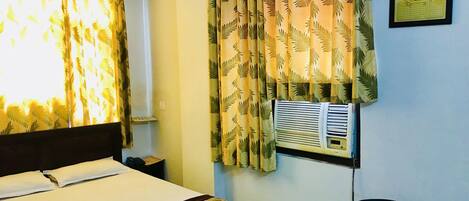 Quarto de hotel com cama, uma cadeira, ar condicionado fixado na parede e cortinas com padrão de folhas.