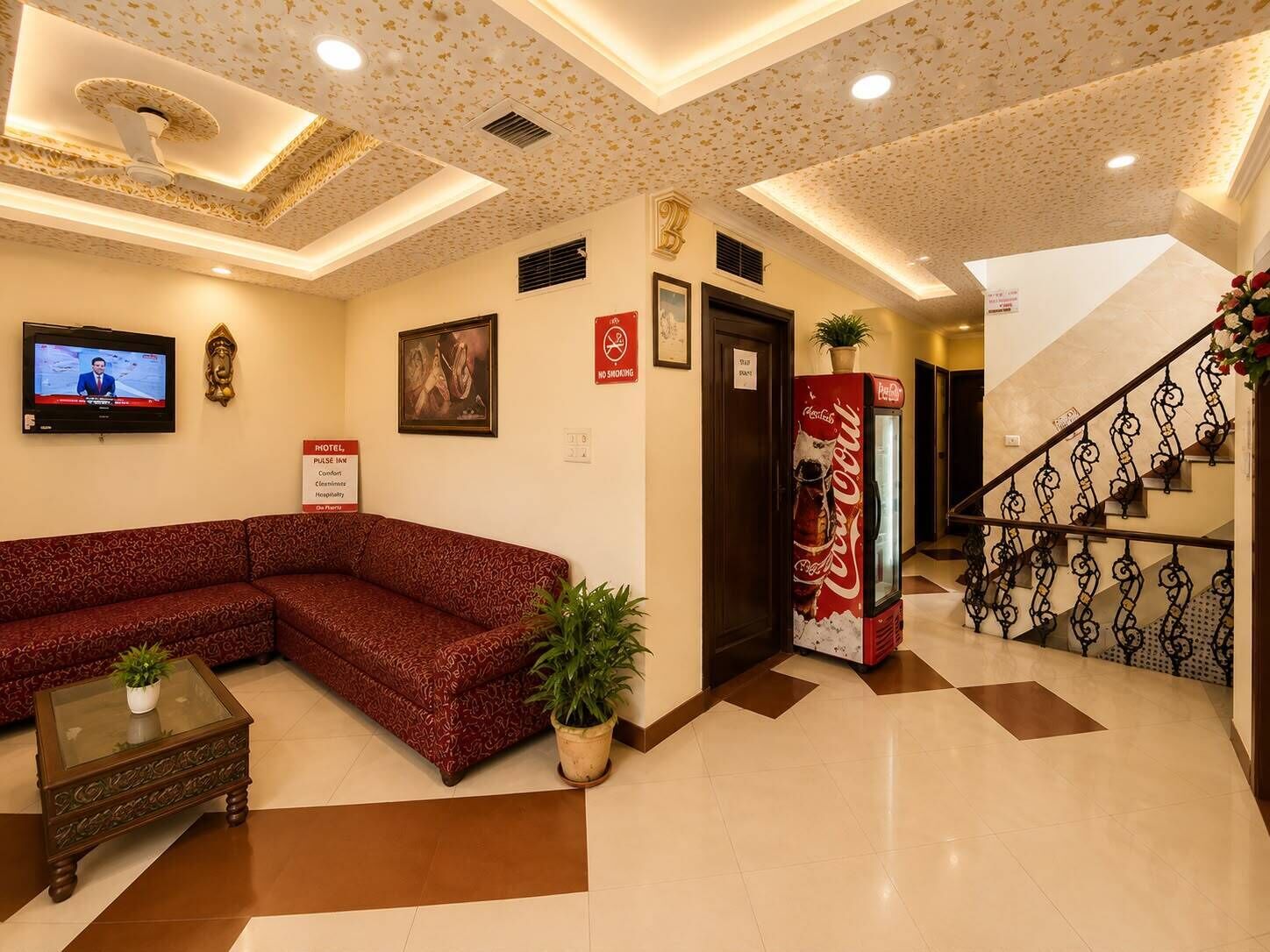 A hotel lobby with a red patterned sofa, a glass-top coffee table, a Coca-Cola refrigerator, a television, and a staircase with a decorative railing.