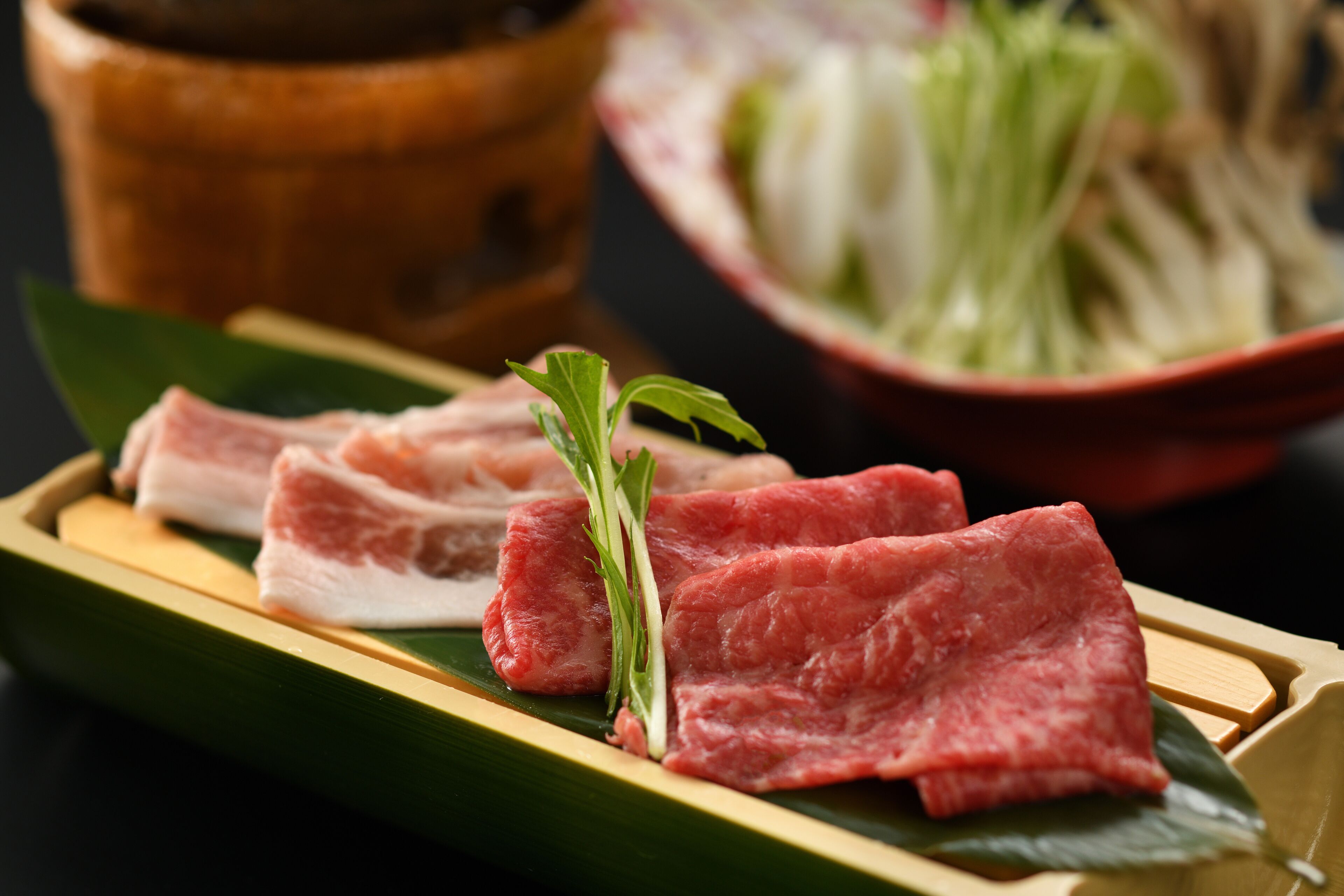A wooden tray with slices of raw meat and green vegetables on a dark surface.