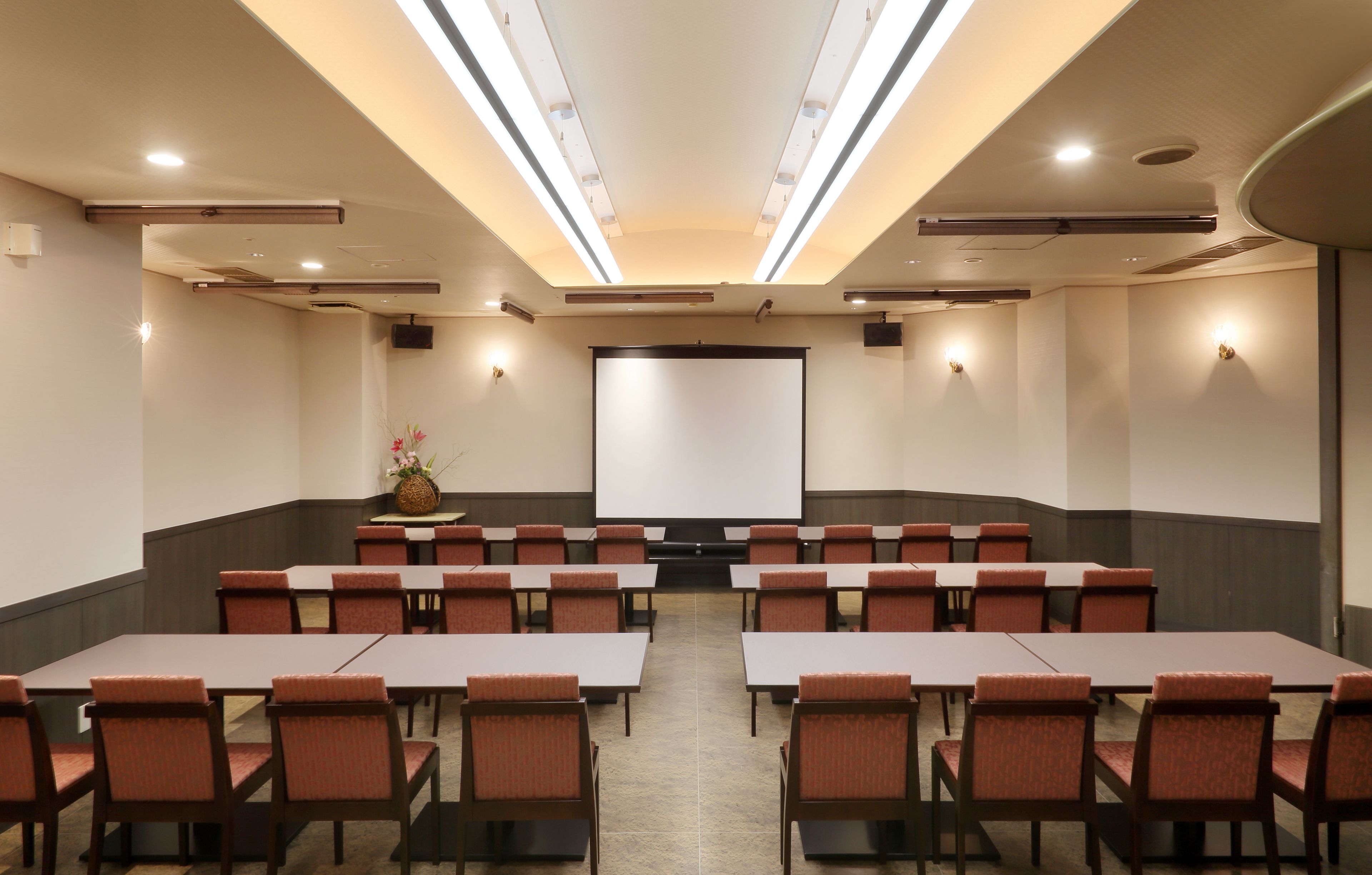 Una sala de conferencias con una pantalla grande, varias mesas y sillas dispuestas para reuniones.