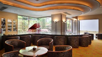 A modern interior with a curved ceiling, seating area with patterned chairs, a bar area with glass cabinets, and large windows offering a view of greenery.