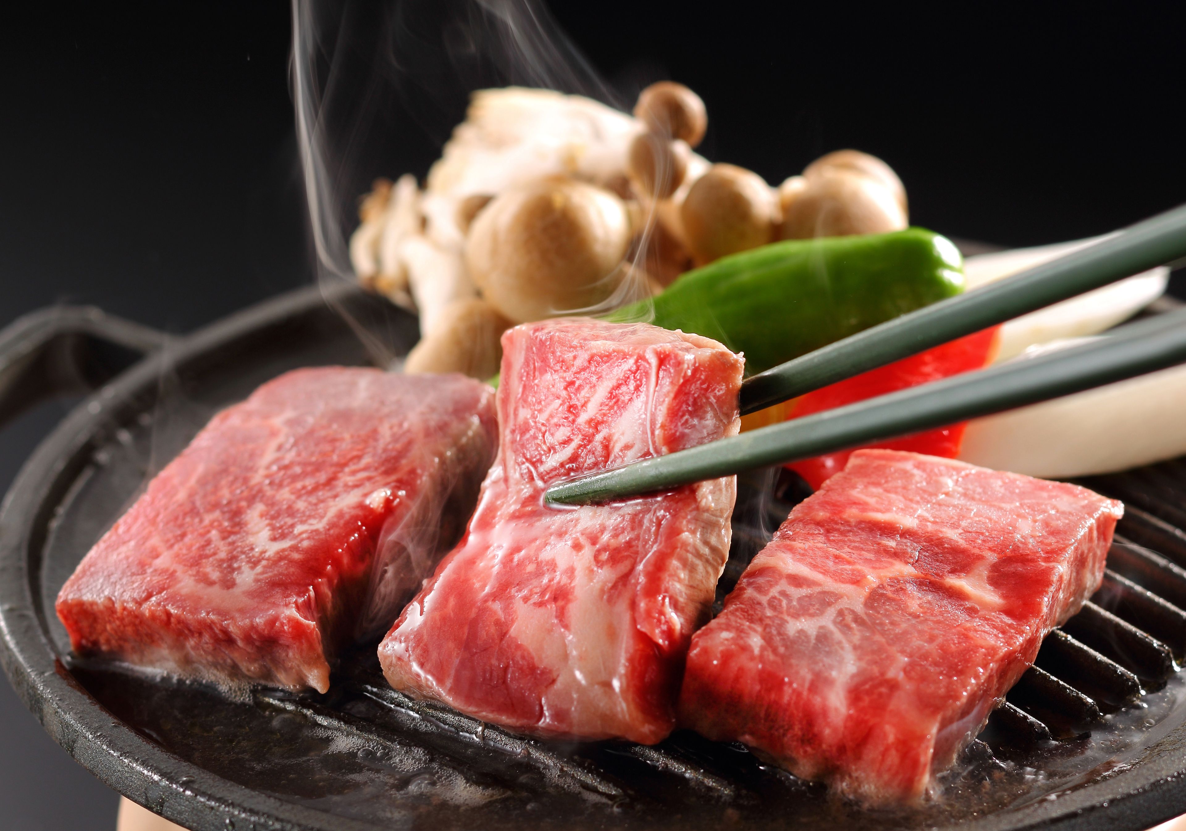 Rohes Fleisch auf einem Grill, dazu Essstäbchen und Gemüse im Hintergrund.