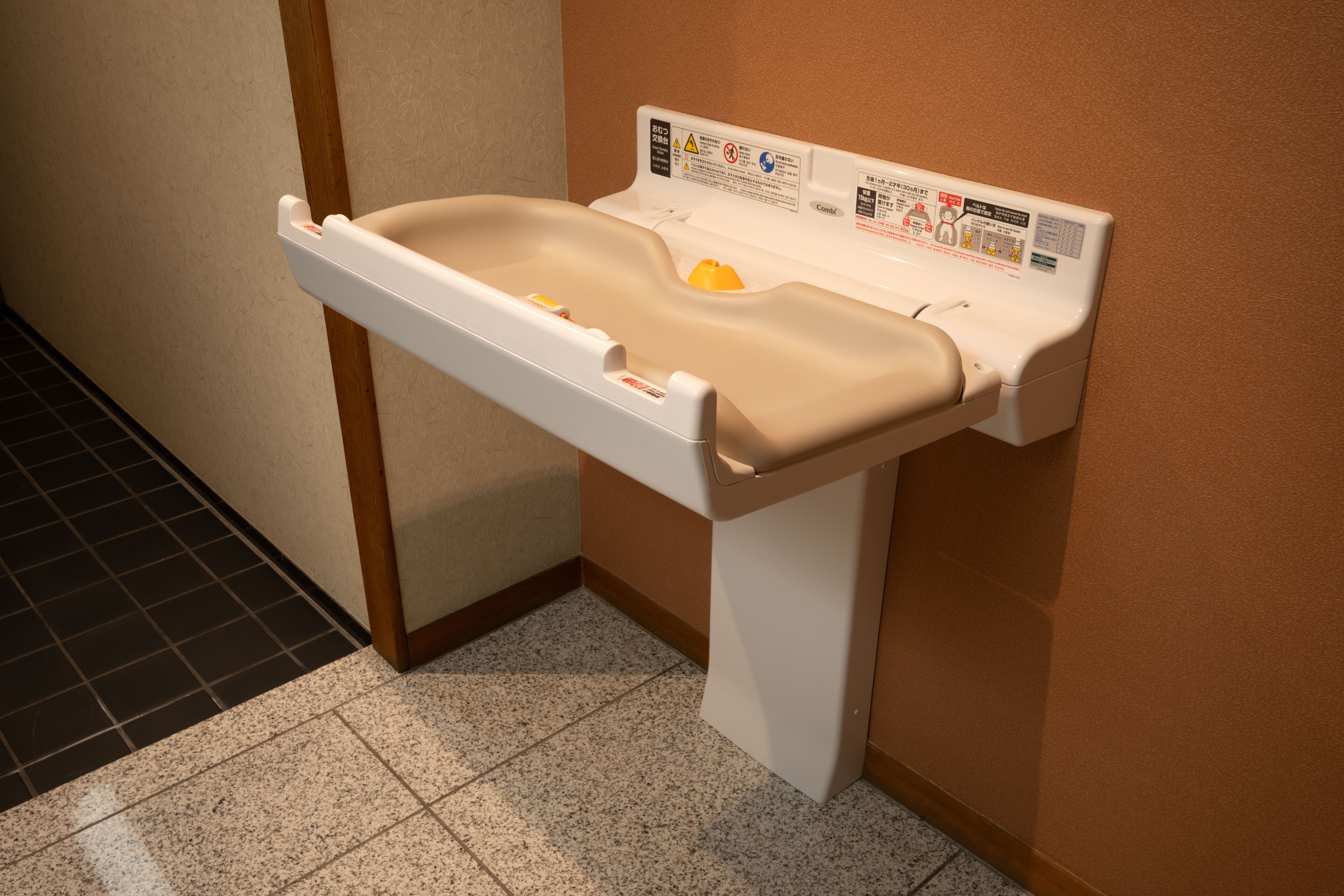 Un lavamanos montado en la pared con un lavabo color beige y un grifo blanco, situado en una esquina con suelo de baldosas y un marco de madera.