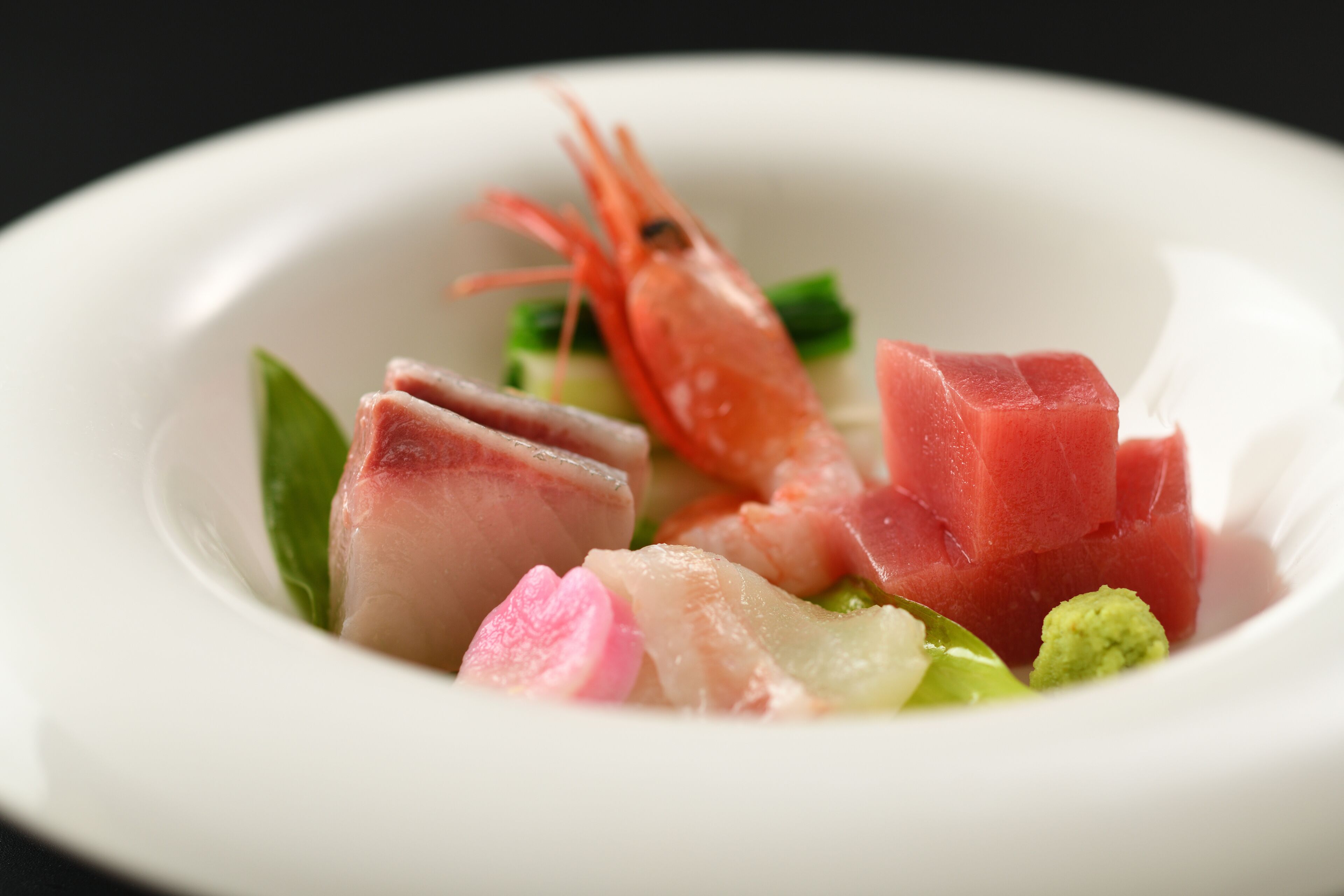 A white plate with a variety of seafood including a shrimp, slices of fish, and some green vegetables.