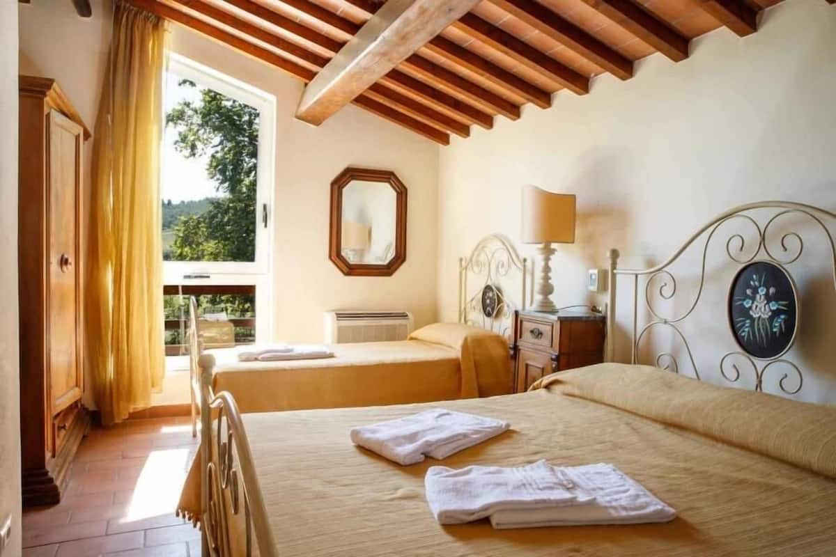 A hotel room with two beds, a wooden ceiling, a window with curtains, a bedside table with a lamp, and a mirror on the wall.