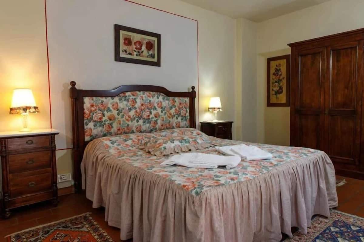 A bedroom with a floral bedspread, wooden headboard, two bedside tables with lamps, a wardrobe, and a patterned rug.