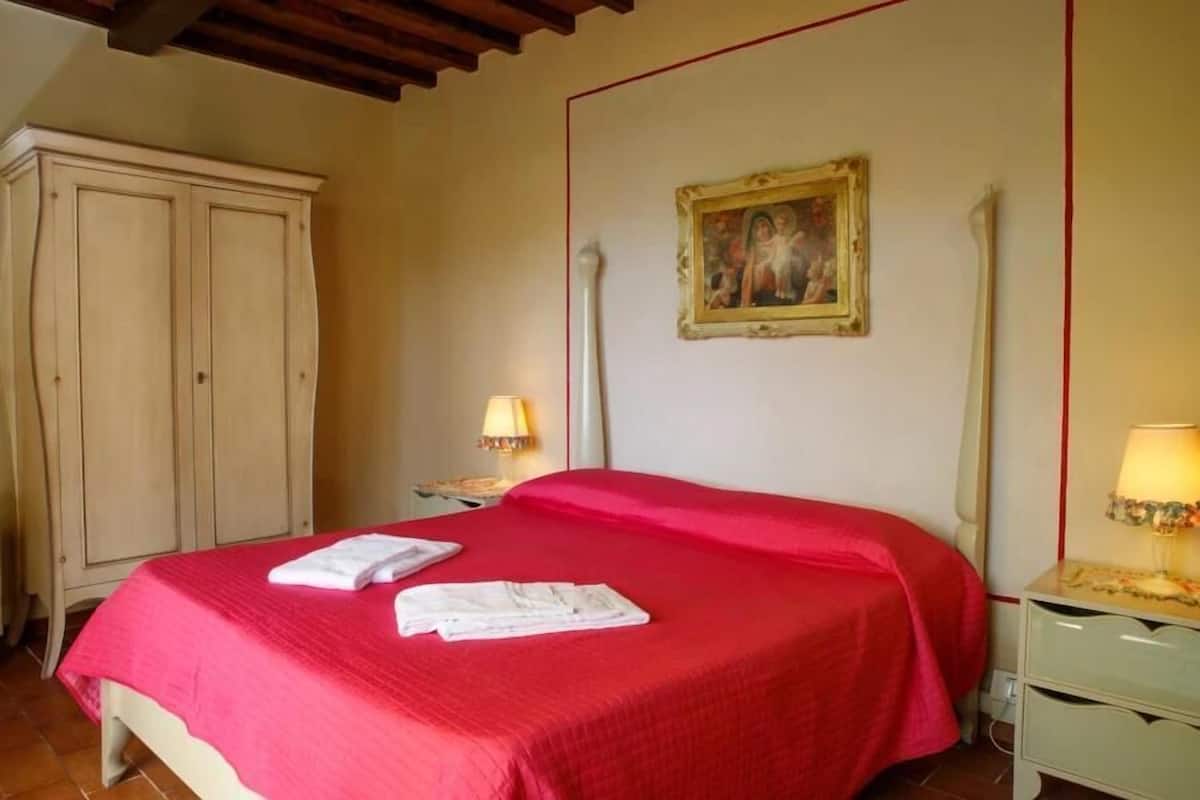 A bedroom with a red bedspread, a wooden wardrobe, a nightstand with a lamp, and a painting on the wall.