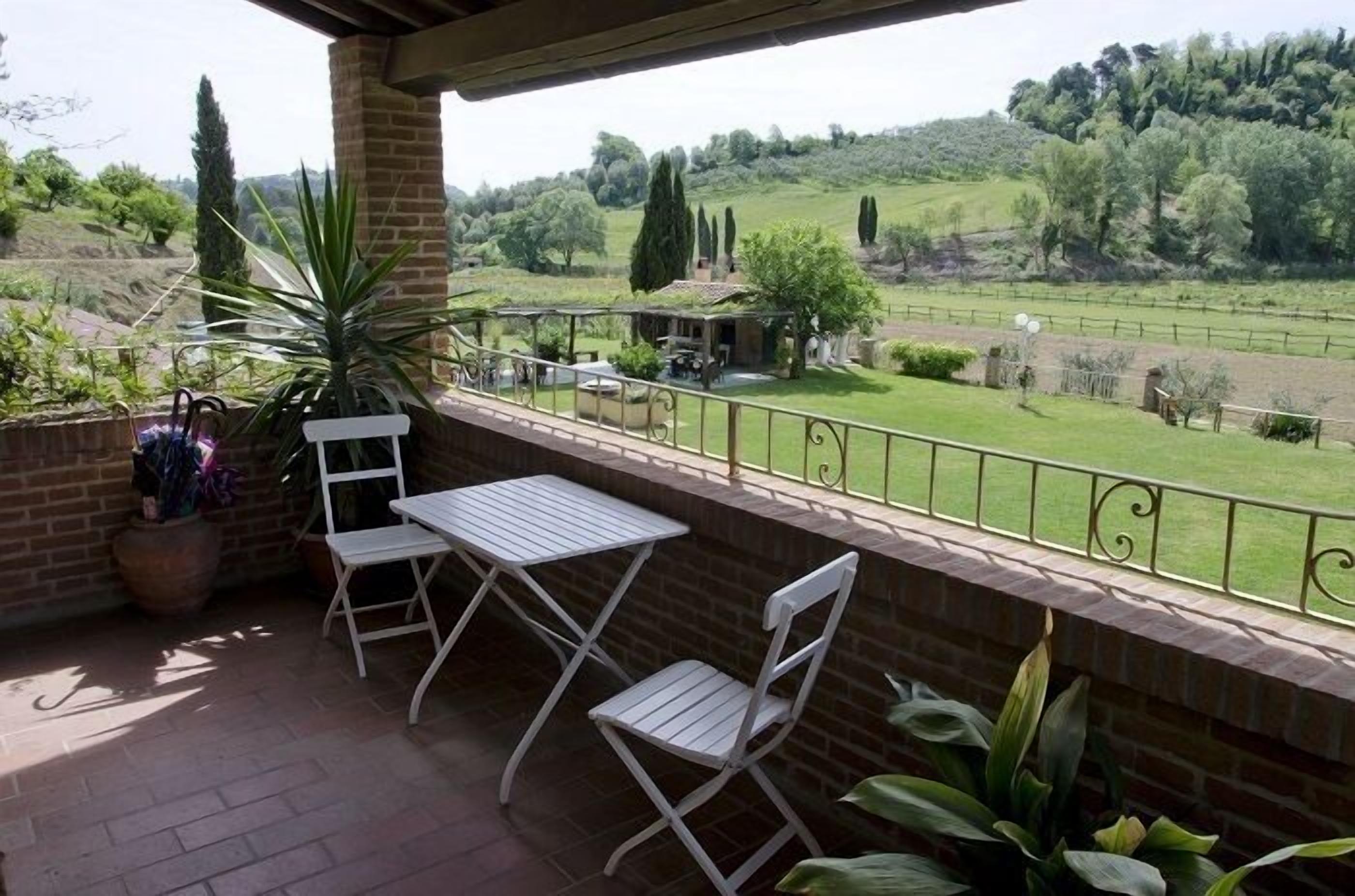 Un balcón con mesa y sillas, con vista a un paisaje verde con árboles y un área cercada.