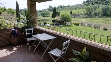 Un balcón con mesa y sillas, con vista a un paisaje verde con árboles y un área cercada.