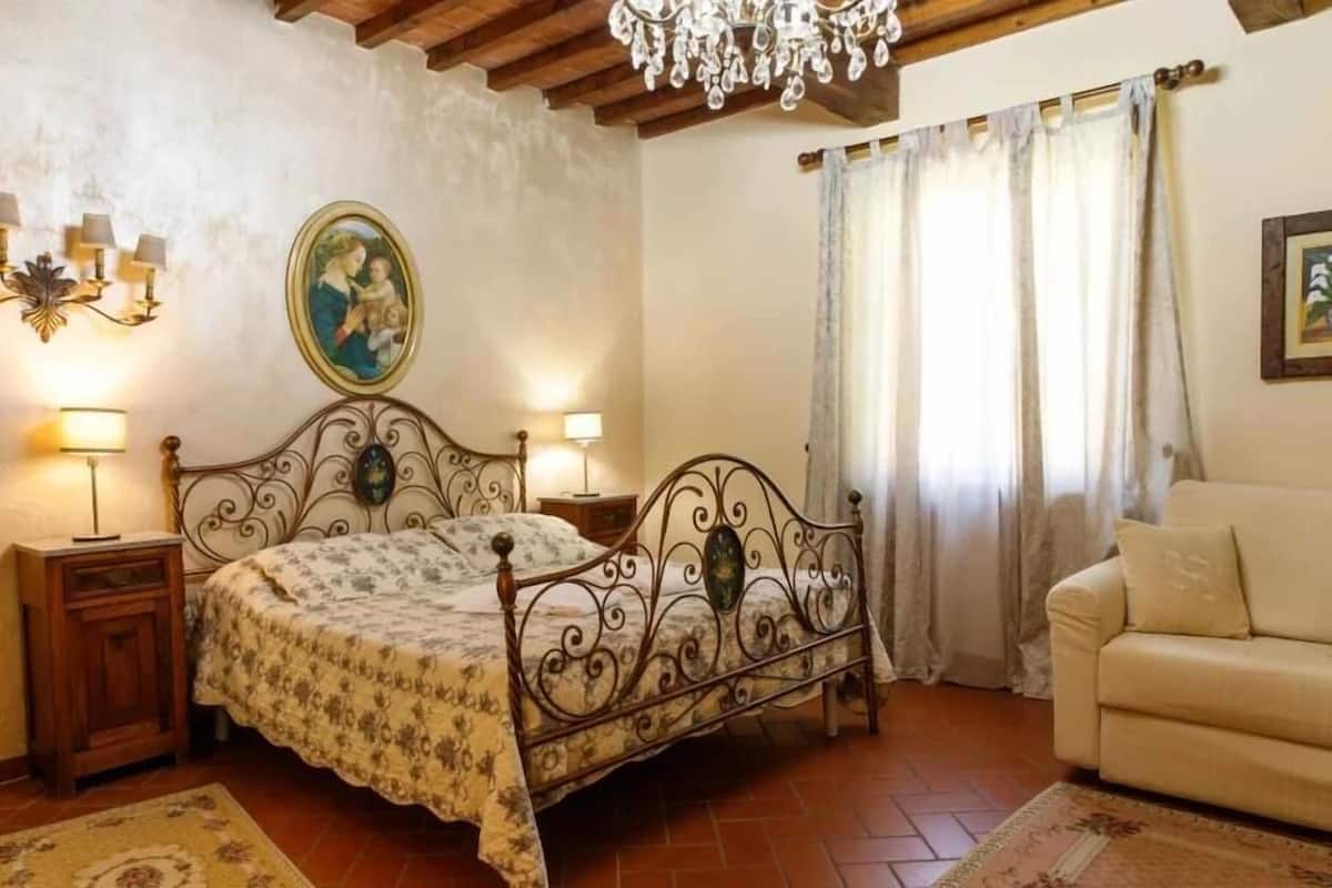 A bedroom with a bed, bedside lamps, a chandelier, a sofa, and a window with curtains.