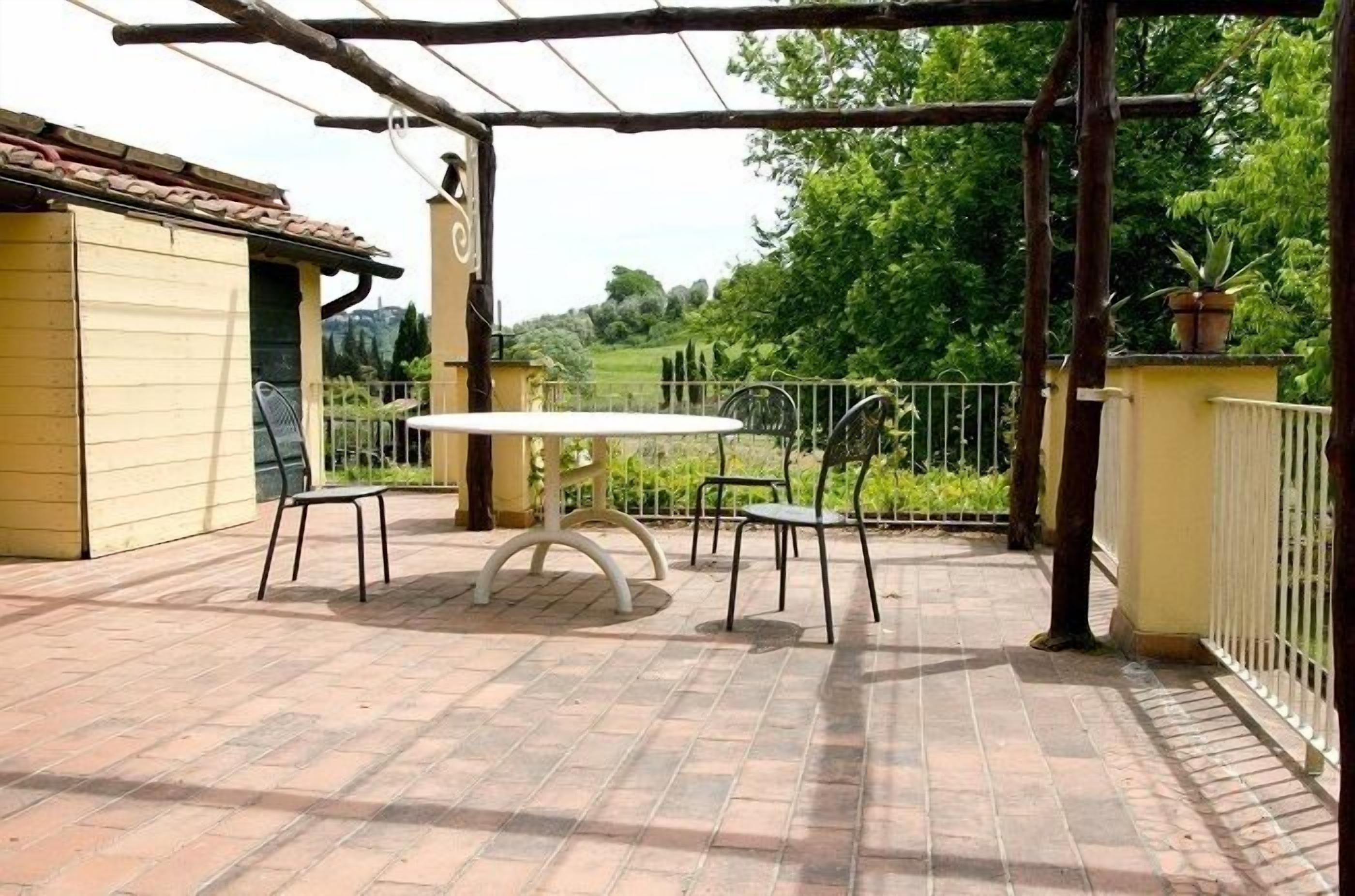 Eine überdachte Terrasse mit rundem Tisch und Stühlen, ein gelbes Gebäude und Blick auf Grünflächen.