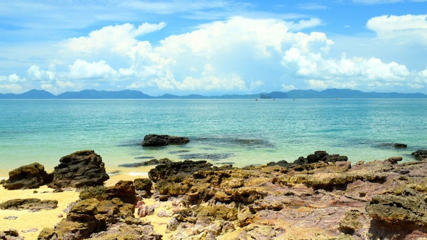A beach with clear turquoise water, rocky foreground, and distant mountains under a partly cloudy sky. - Mukrada House (Krabi)