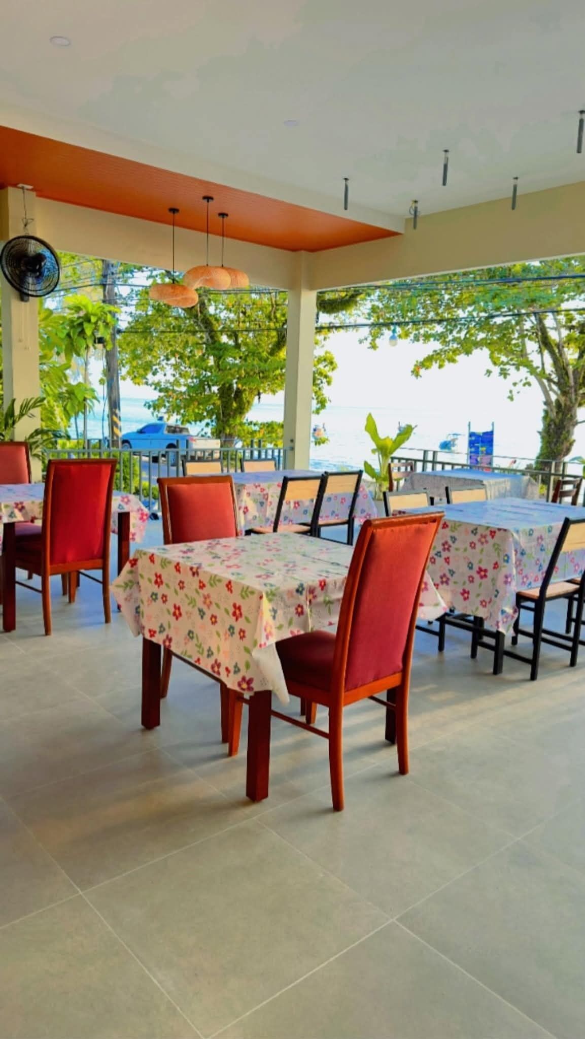 Un comedor interior con mesas y sillas, ventanas amplias con vista al mar y ventiladores de techo.