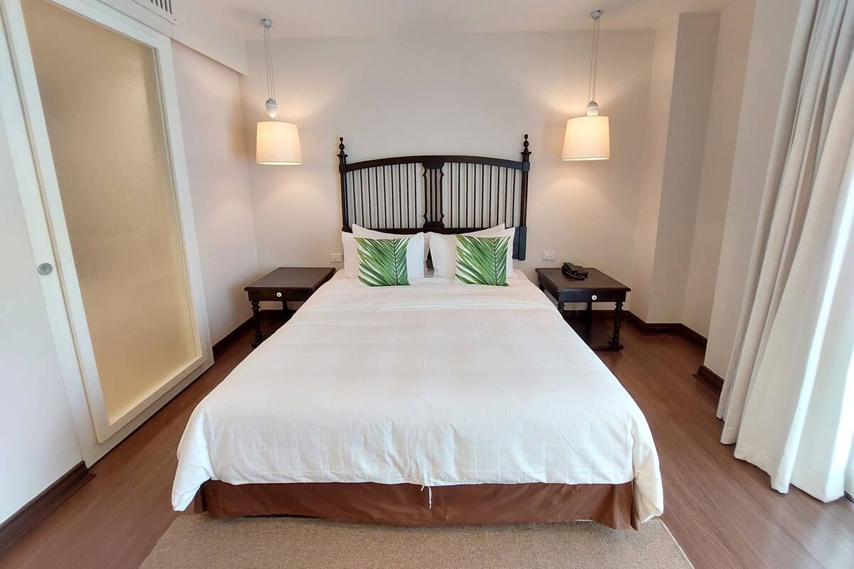 A neatly made bed with white bedding, two bedside tables with lamps, a wooden headboard, and a large wardrobe.