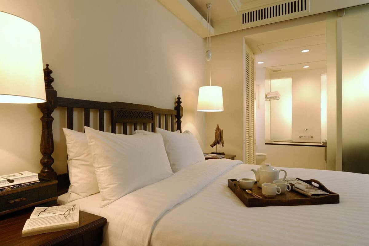 A hotel room with a bed, bedside table with a lamp, a tray with a teapot and cups, and a mirror.