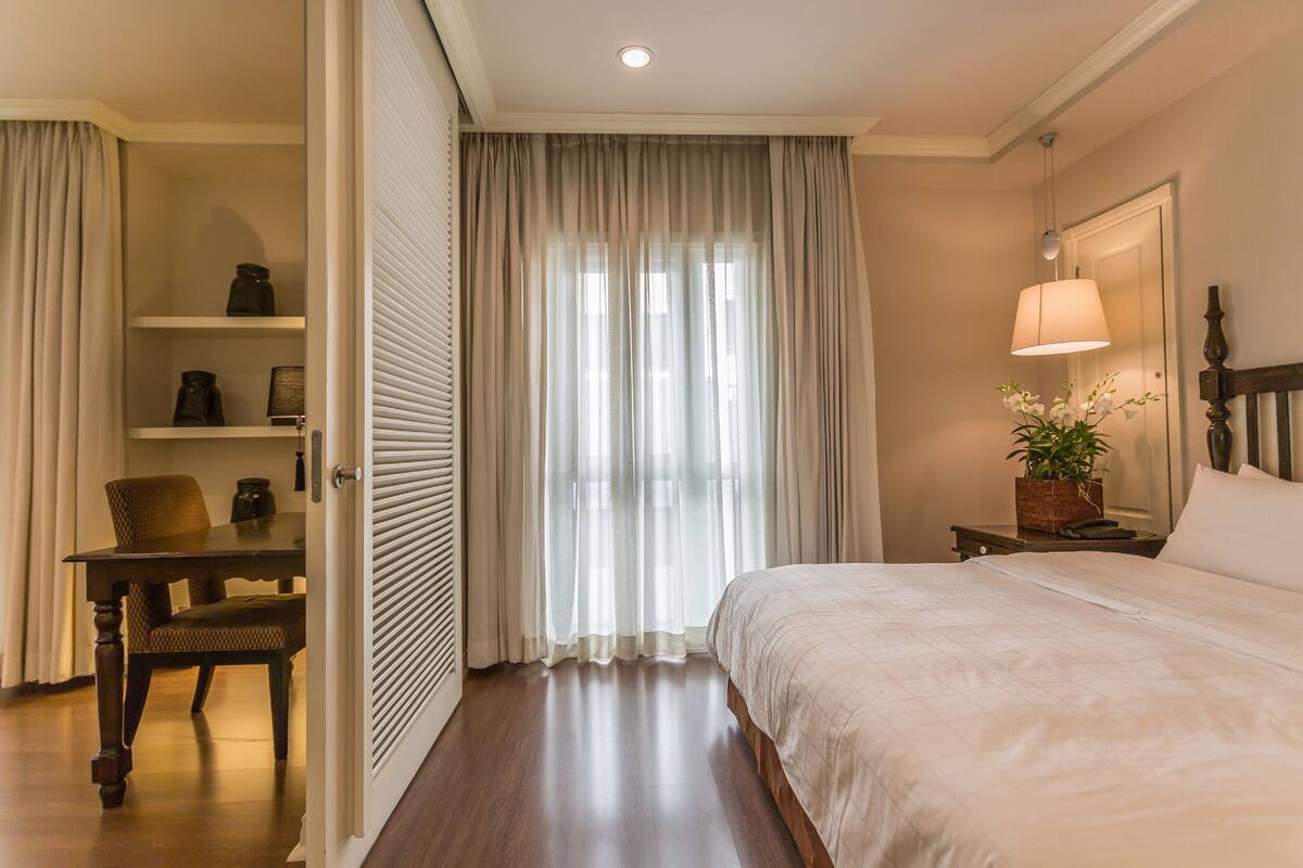A hotel room with a large bed, a desk, a chair, a nightstand with a lamp, and a window with curtains.