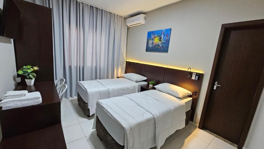 Ein Hotelzimmer mit zwei Betten, einem Schreibtisch mit Stuhl, einer Zimmerpflanze und einem Bild an der Wand.