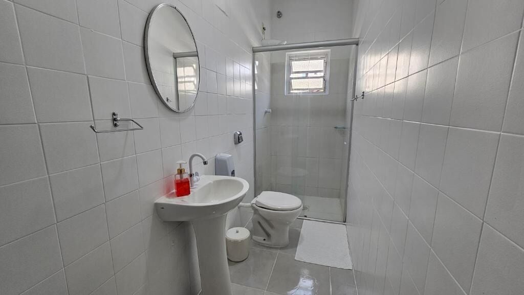 A bathroom with white tiles, a sink, a toilet, a mirror, and a shower area.
