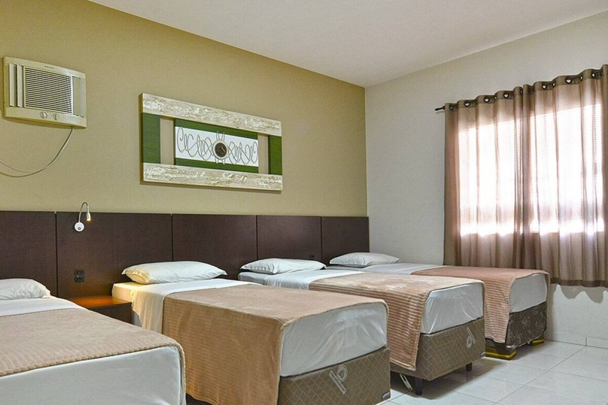 Habitación de hotel con cuatro camas dispuestas en fila, un aire acondicionado de pared, una obra de arte en un marco y una ventana con cortinas.