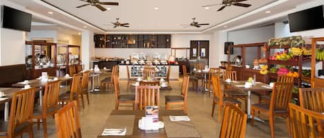 A spacious restaurant with multiple dining tables and chairs, ceiling fans, and a variety of food items displayed on shelves.