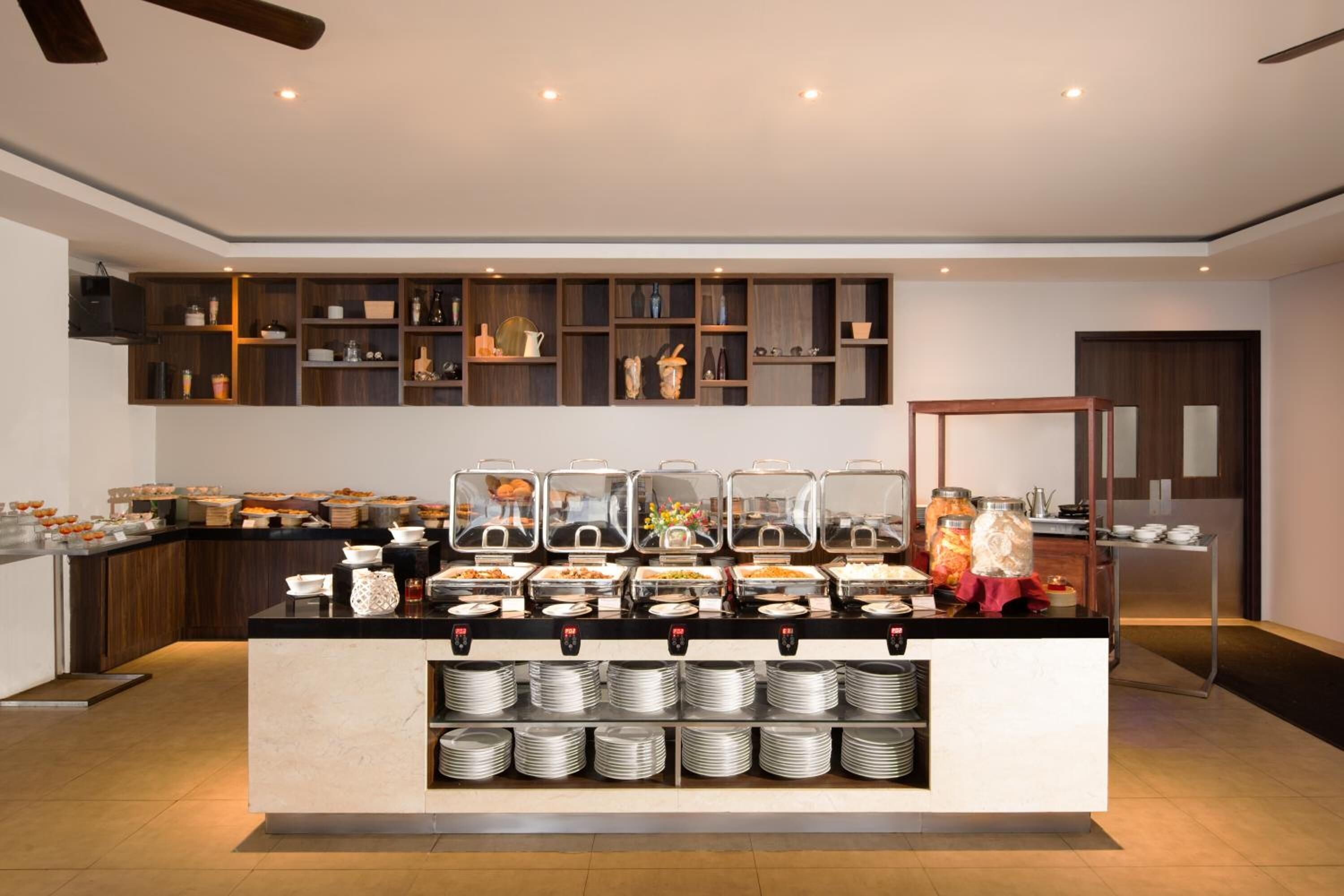 A buffet setup with various food items and containers, a counter with stacked plates, and a shelving unit with kitchenware.
