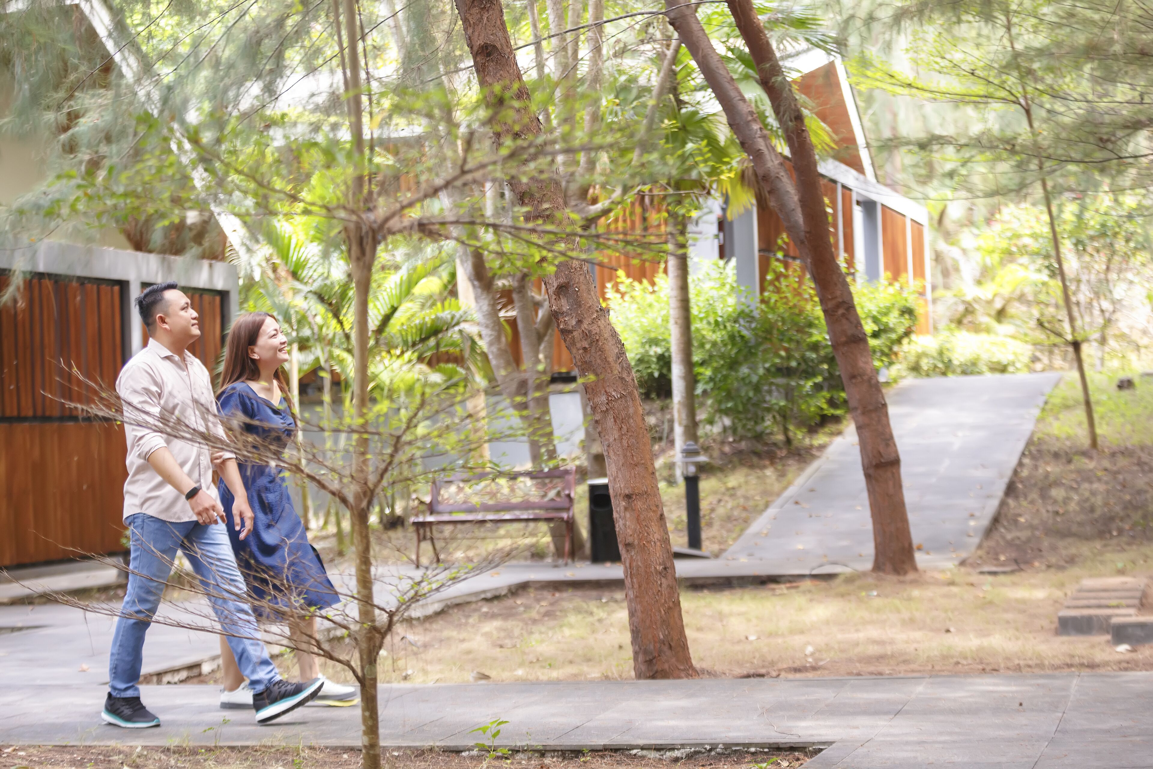 A couple walking on a paved path surrounded by trees and a building with a sloped roof.