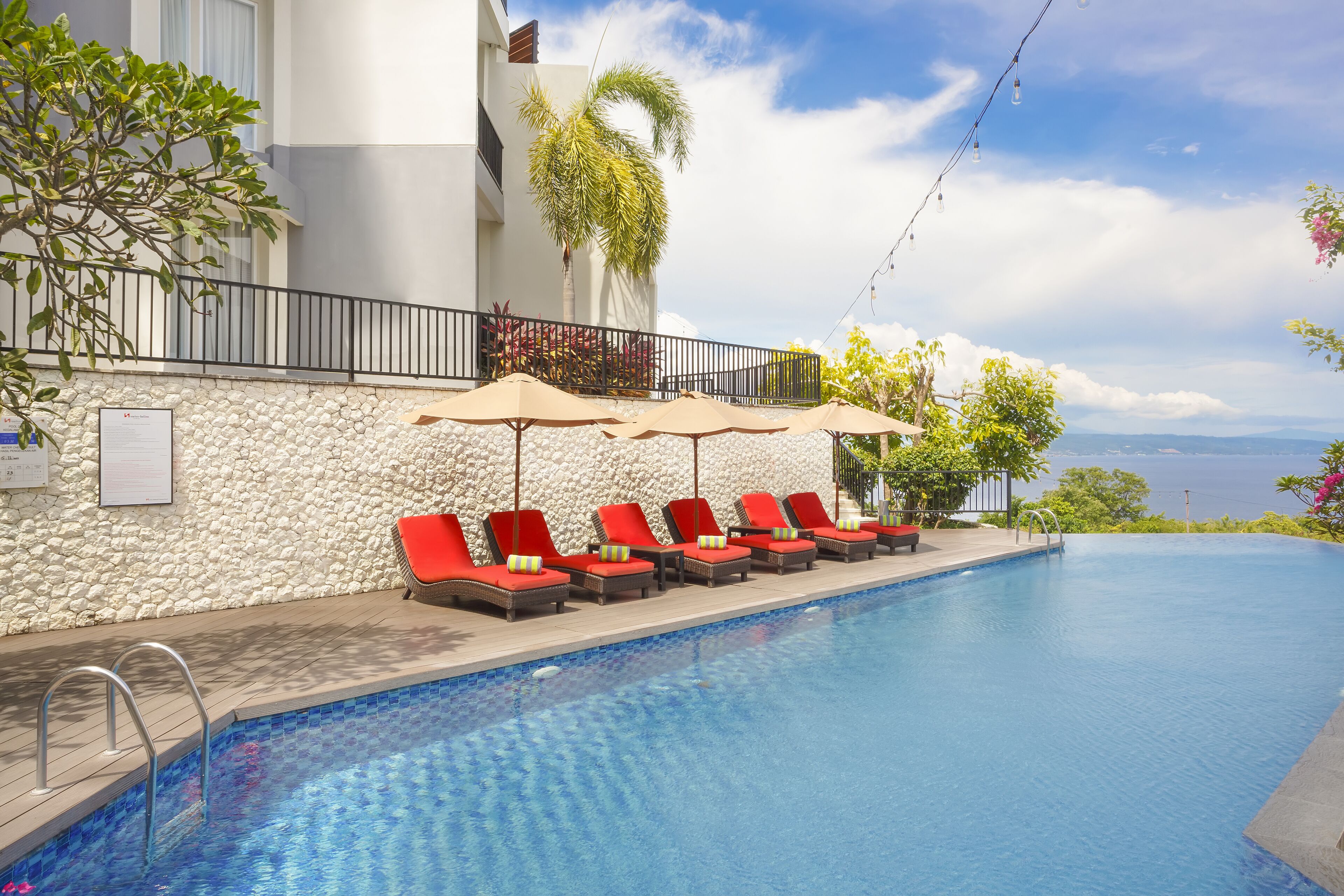 Una piscina con sillas de descanso rojas y sombrillas, un muro de piedra y vistas al mar.