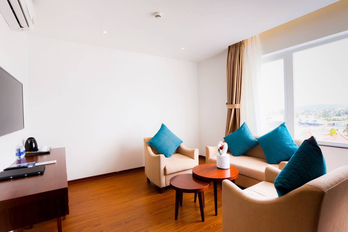 A hotel room with a TV, a desk with a remote, a coffee maker, two sofas with blue cushions, a small table, and a window with a view of a cityscape.